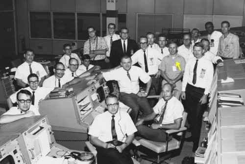 Mission Control team in white shirts and dark ties, gathered around consoles in Houston, mid-1960s.