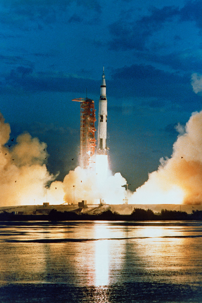 Saturn V rocket lifting off at dawn, reflected across the water, with clouds of fire and smoke at the launch pad.